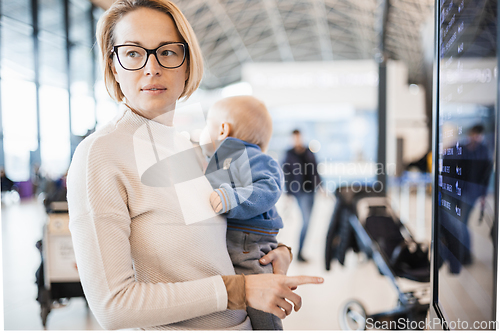 Image of Mother traveling with child, holding his infant baby boy at airport terminal, checking flight schedule, waiting to board a plane. Travel with kids concept.