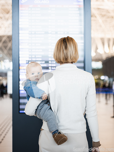 Image of Mother traveling with child, holding his infant baby boy at airport terminal, checking flight schedule, waiting to board a plane. Travel with kids concept.