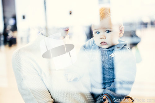 Image of Thoughtful young mother looking trough window holding his infant baby boy child while waiting to board an airplane at airport terminal departure gates. Travel with baby concept.