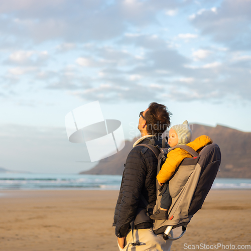 Image of Young father rising hands to the sky while enjoying pure nature carrying his infant baby boy son in backpack on windy sandy beach of Famara, Lanzarote island, Spain at sunset. Family travel concept.