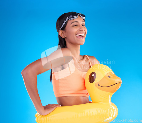 Image of Portrait, wink and rubber duck with a woman on a blue background in studio excited for summer. Happy, smile and playful with an attractive young female ready for swimming on holiday or vacation