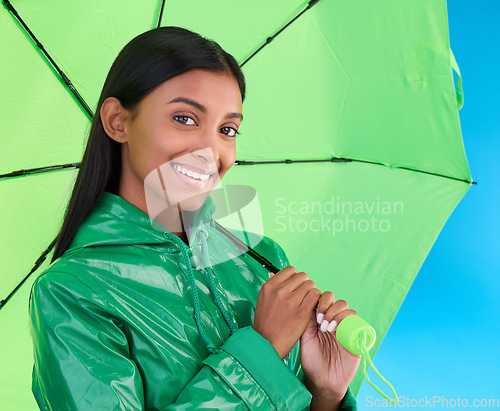 Image of Portrait, smile and female with an umbrella in a studio with a stylish, trendy and fashion green rain coat. Life insurance, face and Indian woman model with winter outfit isolated by blue background.