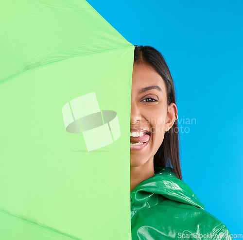 Image of Umbrella, silly and woman portrait with tongue out in studio with raincoat and happiness. Fun, face and playful young female in isolated blue background with comedy, emoji and funny joke with a smile