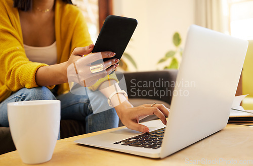 Image of Closeup of woman working remote while typing on her laptop and holding her smartphone sitting on a sofa in a bright living room. One focused hispanic young female at home using modern technology