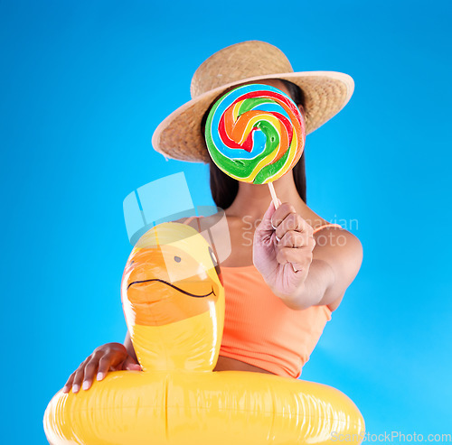 Image of Pool float, woman and lollipop in a studio with sweet snack and swimsuit with food. Isolated, blue background and holiday outfit of a young female hiding with sweets, candy or rubber duck feeling fun