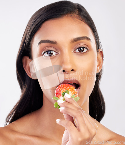 Image of Portrait, beauty and a woman biting a strawberry in studio on a gray background for health, diet or nutrition. Face, skincare and serious with an attractive young female model eating a fruit berry