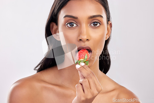 Image of Portrait, skincare and a woman biting a strawberry in studio on a gray background for health, diet or nutrition. Face, beauty and serious with an attractive young female model eating a fruit berry