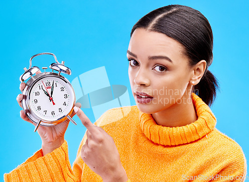 Image of Woman, annoyed and pointing at alarm clock in portrait for warning by blue background in studio. Gen z girl, student or model with watch, time management and schedule with angry face to start morning