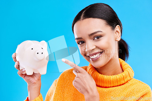 Image of Happy woman, money and pointing to piggy bank for investment, budget or finance against blue studio background. Portrait of female with savings, cash or coin for profit investing or financial freedom