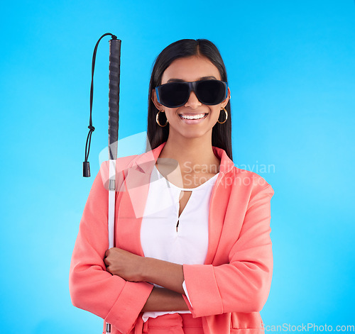 Image of Blind, disability and sunglasses with a woman on a blue background in studio holding her walking stick. Portrait, vision and smile with an attractive young female arms crossed for disabled lifestyle