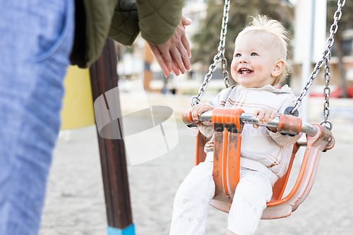 Image of Mother pushing her cheerful infant baby boy child on a swing on sandy beach playground outdoors on nice sunny cold winter day in Malaga, Spain.