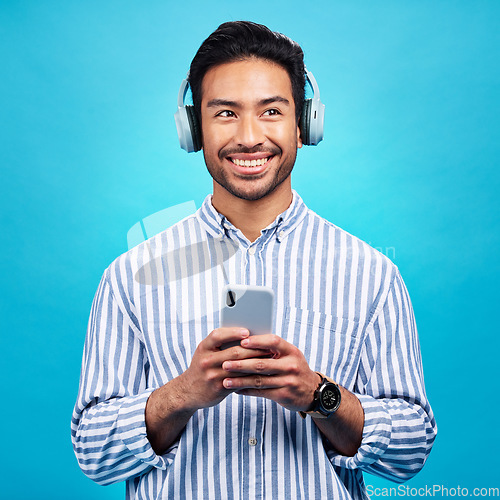 Image of Headphones, music and thinking man with phone in studio for streaming, audio and subscription against blue background. Radio, smile and happy asian male online for podcast, track and internet app