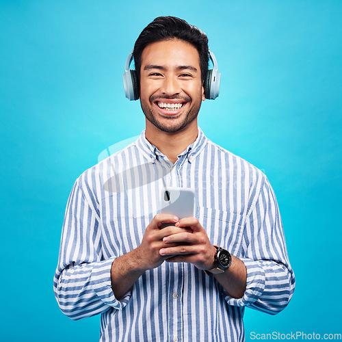 Image of Music, headphones and happy man with phone in studio for streaming, audio and subscription against blue background. Radio, smile and face of asian male online for podcast, track and internet app