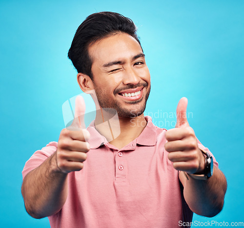 Image of Portrait, thumbs up or wink by man in studio in positive, feedback or review on blue background. Face, smile and hand gesture by Asian male model showing yes, agreement or vote emoji while isolated