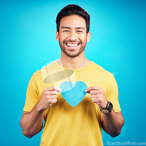 Image of Portrait, cutout heart and Asian man with happiness, loving and emoji with guy against a blue studio background. Face, male person and model with symbol for love, smile and carefree with support