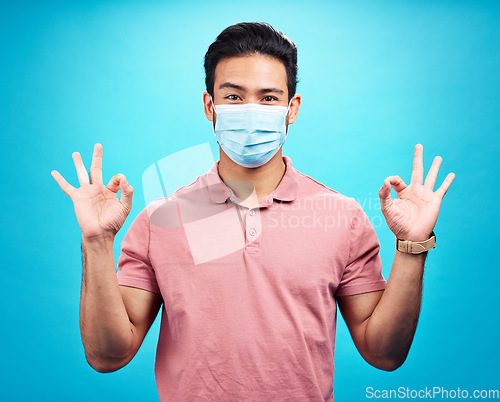 Image of Man in face mask, portrait and covid with okay hand gesture, safety from virus and bacteria isolated on blue background. Disease, corona and male person with emoji and health compliance in studio