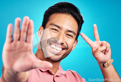 Image of Happy asian man, portrait and peace sign for selfie, profile picture, or social media against a blue studio background. Male influencer or vlogger with smile showing peaceful emoji for photo or vlog