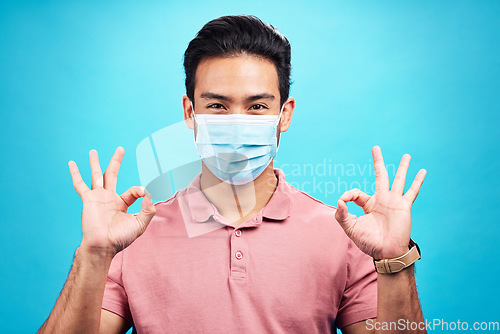 Image of Man in face mask, portrait and corona with okay hand sign, safety from virus and bacteria isolated on blue background. Disease, covid and male person with emoji and health compliance in studio