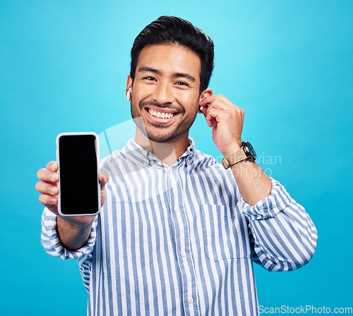 Image of Asian man, phone and mockup screen with earphones for music listening, call or audio against a blue studio background. Portrait of happy male showing smartphone display for sound app or communication