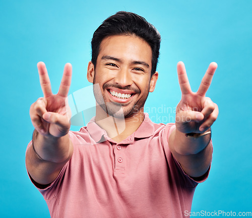 Image of Portrait, peace sign and Asian man with a smile, support and confident on a blue studio background. Face, male person or model with v gesture, emoji and symbol with happiness, hands and peaceful icon