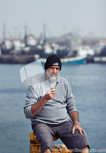 Image of Fisherman, mature man portrait and smoking pipe by the sea water at a port for fishing. Old man, ocean and senior dock worker sitting by boats outdoor with tobacco smoke at a harbor for work