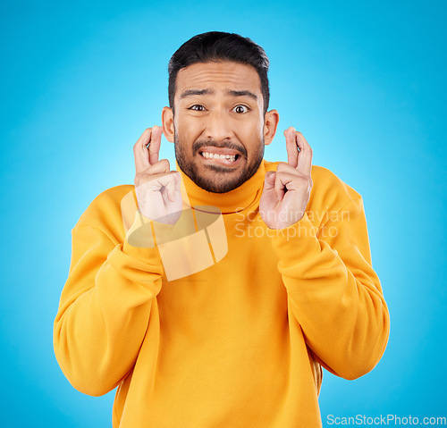 Image of Nervous, portrait and fingers crossed by asian man with anxiety in studio for news, feedback or review on blue background. Hand, emoji and face of guy with stress for results, competition or giveaway