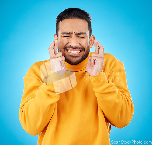 Image of Nervous, eyes closed and fingers crossed by asian man with anxiety in studio for news, feedback or review on blue background. Hand, emoji and guy with stress for results, competition or giveaway