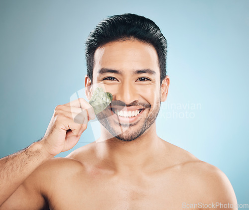 Image of Face, man and beauty, gua sha and cosmetic tools, smile in portrait and natural treatment on blue background. Skincare, healthy glow and jade stone, dermatology and wellness with male model in studio