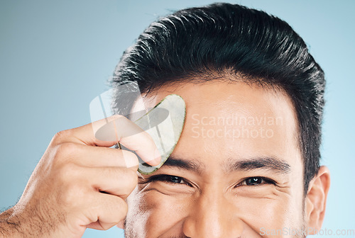 Image of Closeup, man and beauty, gua sha and cosmetic tools, eyes and portrait with natural treatment on blue background. Skincare, glow and jade stone, dermatology and wellness with male model in studio