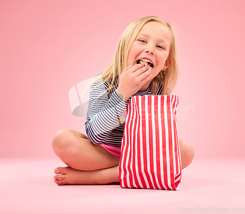 Image of Popcorn eating, portrait and happy girl in a studio with pink background sitting with movie snacks. Food taste, happiness and hungry young child with a paper bag and chips feeling relax with a smile