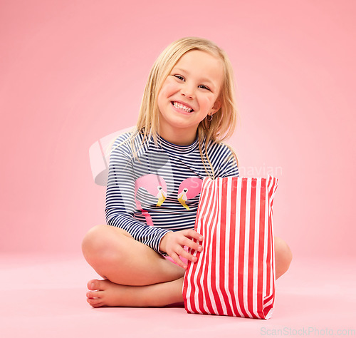 Image of Popcorn, food and happy girl portrait in a studio with pink background sitting with movie snacks. Snack, happiness and hungry child with a paper bag and chips eating and feeling relax with a smile