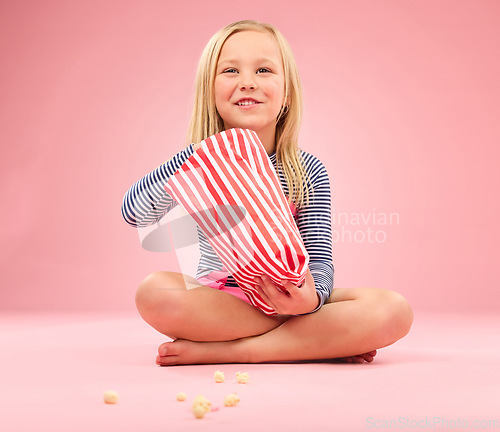 Image of Popcorn, snack and happy girl in a studio with pink background sitting with movie snacks. Food, happiness and hungry young child with a paper bag and chips eating and feeling relax with a smile