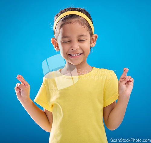 Image of Child, hand and fingers crossed in studio for hope, wish or good luck against a blue background. Girl, eyes closed and emoji hands, praying and wishing, happy and smile while posing on isolated space