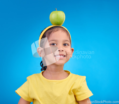 Image of Portrait, smile and girl with apple on head on blue background for nutrition, healthy eating and diet. Food, youth and face of happy young girl in studio with fruit for organic, vitamins and wellness