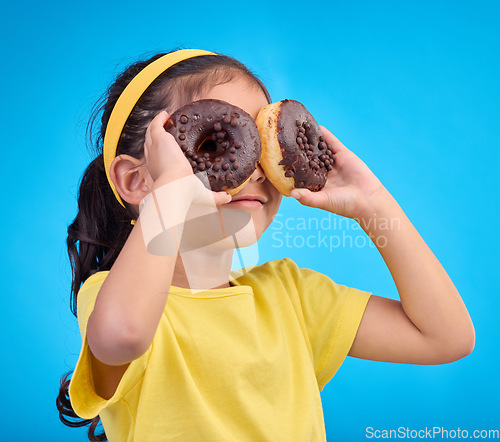 Image of Doughnuts, eyes and face of playful child with food isolated against a studio blue background with a smile. Adorable, happy and cute young girl or kid excited for sweet sugar donut calories