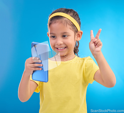 Image of Phone, girl kid and peace sign selfie in studio isolated on a blue background. Technology, v hand emoji and smile of child with mobile smartphone to take pictures for happy memory or social media.