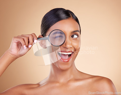 Image of Face, beauty or magnifying glass with a model woman in studio on beige background searching for skincare. Facial, eye wink or excited Indian girl looking for luxury self care cosmetics or makeup