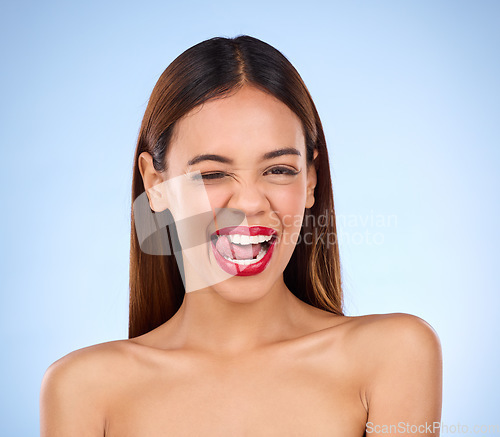 Image of Wink, woman and red lipstick makeup portrait with cosmetics on face in studio. Aesthetic female model on a blue background for self care, facial glow and beauty or color for skin with tongue out