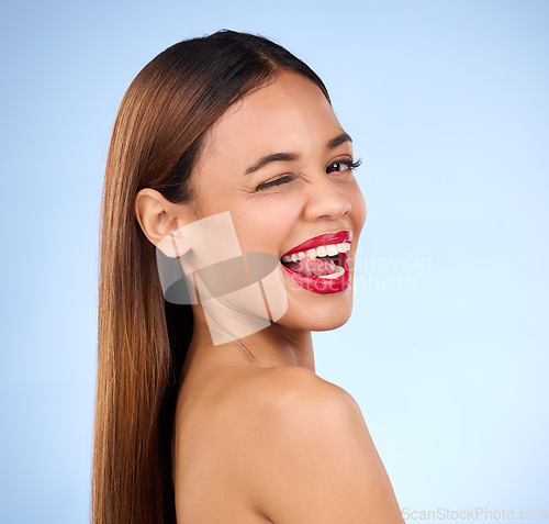 Image of Beauty, woman and wink for face makeup and skincare cosmetics in studio. Female model person with tongue on teeth and red lips on a blue background for self care, facial glow and healthy skin shine