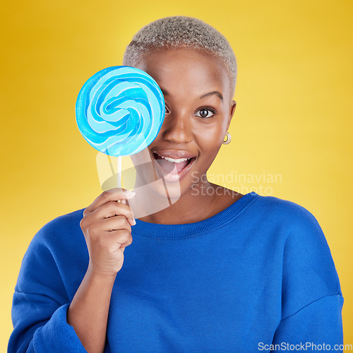 Image of Excited, portrait and black woman with a lollipop in studio for dessert, sweet treat or craving. Happy, surprise and face of African female model posing with blue candy isolated by yellow background.