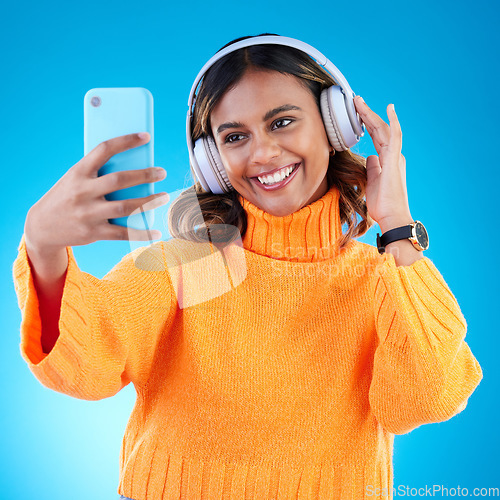 Image of Music headphones, smile and selfie of woman in studio isolated on a blue background. Profile picture, radio and happy Indian female streaming, listening and enjoying sound, audio podcast or song.