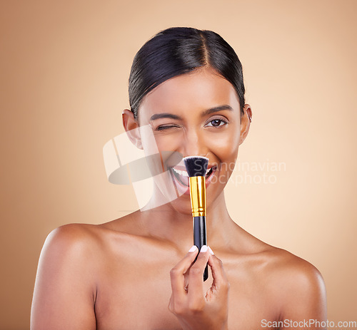 Image of Wink, brush or makeup girl with beauty, cosmetic products or self care on studio background. Flirt, happy model face or young Indian woman with luxury cosmetics, glowing skincare or facial smile