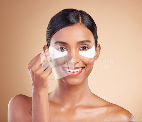 Image of Eye patch, beauty and portrait of a woman in studio for skincare, dermatology and cosmetics. Happy Indian female model with smile and collagen gel mask for self care facial on brown background