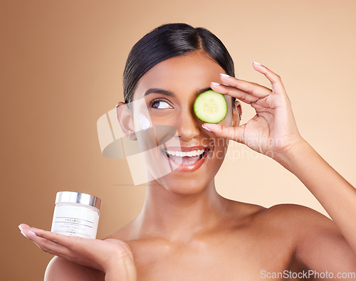 Image of Beauty, skincare and Indian woman with cucumber, cream and dermatology against a brown studio background. Female, cosmetics and happy lady with natural care, lotion and healthy detox with grooming