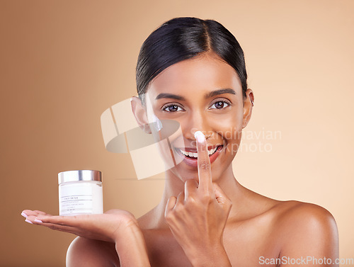 Image of Portrait, product and nose with a model woman in studio on a beige background to apply facial lotion. Face, beauty and skincare with an attractive young female applying moisturizer to her skin