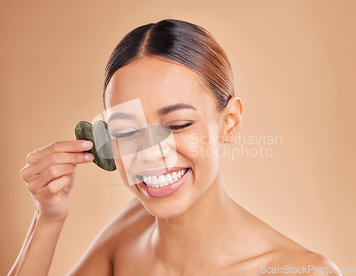 Image of Happy, facial and massage with gua sha by woman in studio for skincare, cosmetics and facelift on brown background. Smile, face and anti aging tool for girl beauty model relax with luxury dermatology