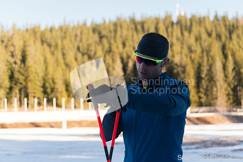 Image of Handsome male athlete with cross country skis, taking fresh breath and having break after hard workout training in a snowy forest. Checking smartwatch. Healthy winter lifestyle.