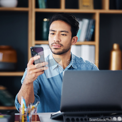 Image of Serious asian man, phone and laptop for social media, communication or networking at office desk. Male creative designer looking at smartphone by computer for browsing, research or planning startup