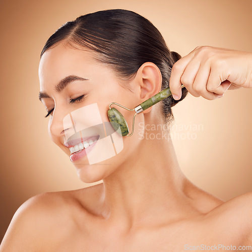 Image of Skincare, jade roller and happy woman in studio for beauty, wellness and anti aging against brown background. Facial, skin and girl relax with massaging tool, facelift and lymphatic drainage isolated