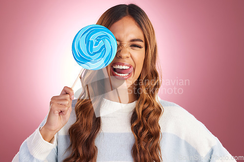 Image of Portrait, funny face and lollipop with a woman in studio on a pink background holding giant candy. Comic, comedy and sweet with a playful young female feeling silly while eating a blue sugar treat